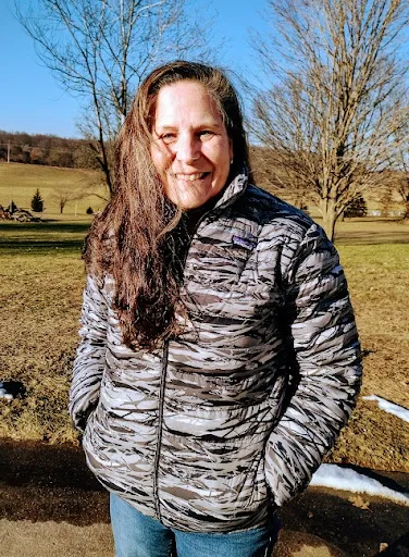 Nancy smiling outdoors on a sunny winter day in a patterned puffer jacket, with open fields and bare trees behind her.